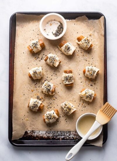 pigs in a blanket on a baking sheet with melted butter, pastry brush, and bagel seasoning prior to baking. 