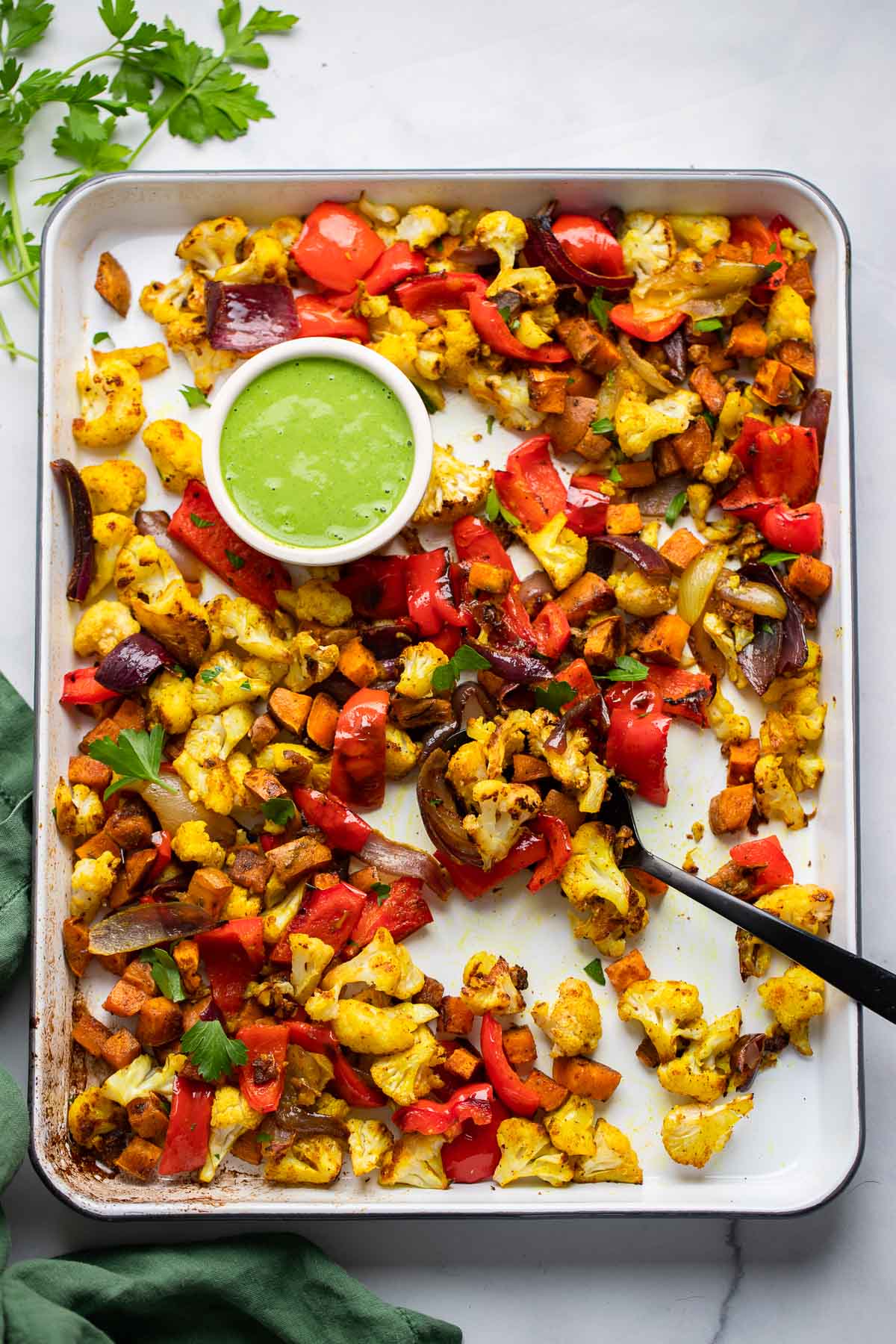 curry roasted vegetables in a sheet pan with a green yogurt sauce and spoon for serving.